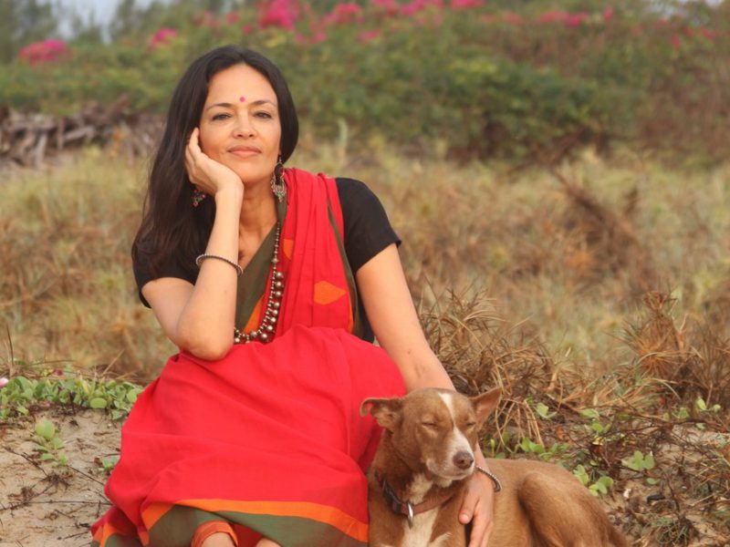 Photograph of a woman with long hair in red sari, sitting with a brown dog against a background of bushes with vines and pink azaleas. The woman is resting her chin on one hand and petting the dog with the other.