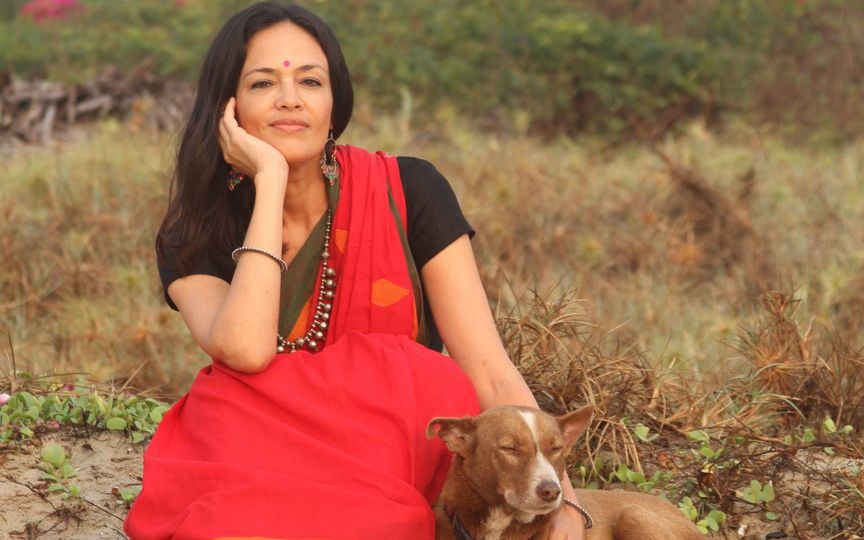 Photograph of a woman with long hair in red sari, sitting with a brown dog against a background of bushes with vines and pink azaleas. The woman is resting her chin on one hand and petting the dog with the other.