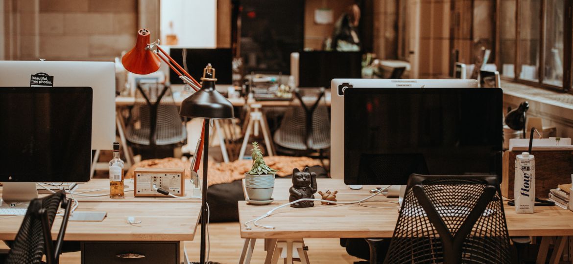 An office space with rows of desks. At the forefront are two desks with monitors, chairs and desk lamps. There are knick-knacks like a wooden statue of a cat, plan, a bottle containing amber-coloured liquid and cables.