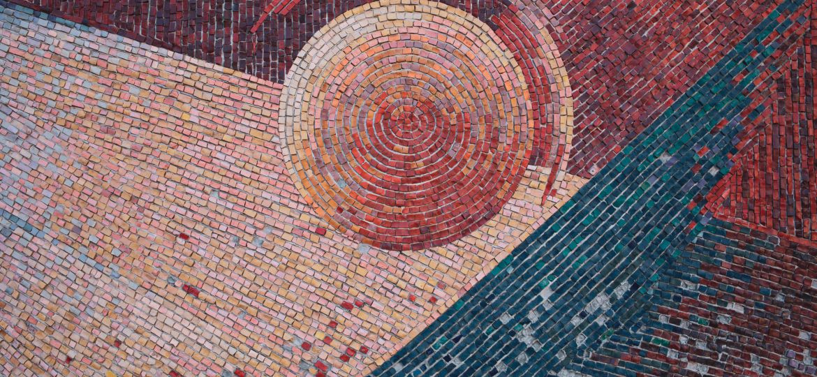 A photograph of a mosaic pattern in hues of red and one streak of green. At the centre of the pattern is a circle surrounded by a semi circle.