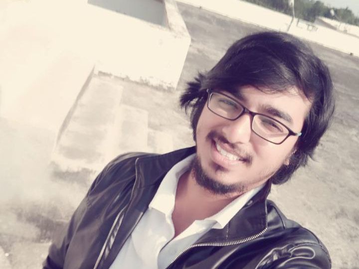 Photo of a young smiling man taken from an upper angle. He has side-swept black hair and a bear. He is wearing a white shirt, black leather jacket and square spectacles.
