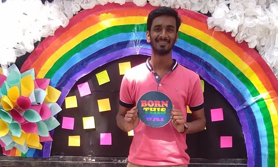 photo of Rajeev in a pink tee-shirt and light brown pants and holding a sign saying 'Born this way'. They are standing in front of a pride rainbow pasted on a wall. There are colourful post-its on a black patch on the wall.