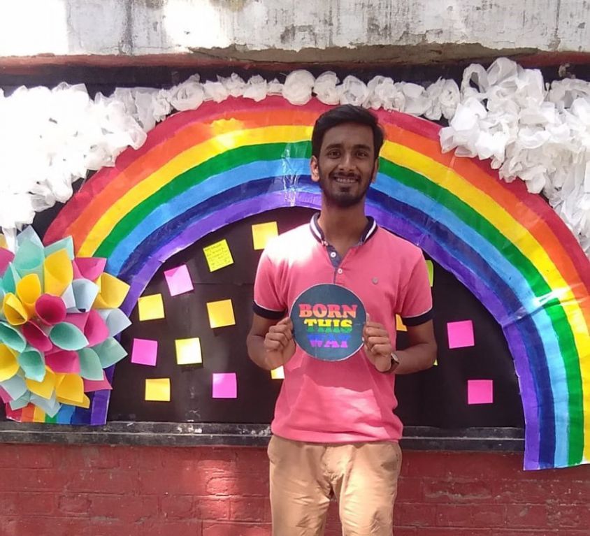 photo of Rajeev in a pink tee-shirt and light brown pants and holding a sign saying 'Born this way'. They are standing in front of a pride rainbow pasted on a wall. There are colourful post-its on a black patch on the wall.