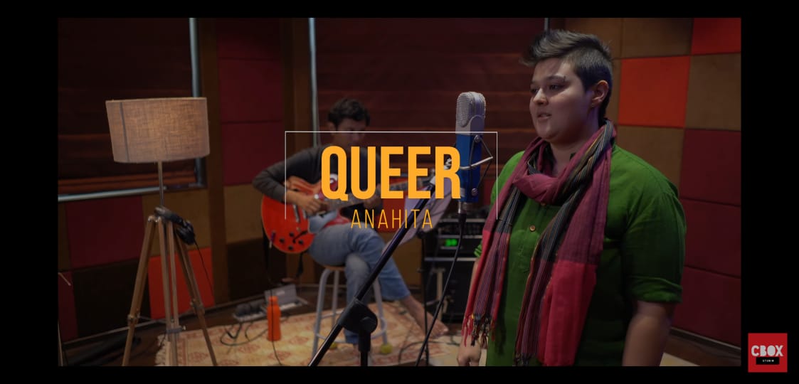 Screenshot from the video, with Anahita standing in front of a mic, wearing a green top and a colourful scarf around the neck. Behind Anahita is a guitar player seated on a stool. To the right of the guitar player is a lamp on a stand