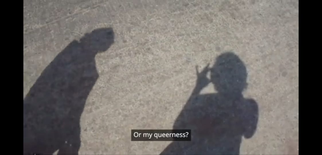 the image is a screenshot from the video and has the shadow of Sonali Udaybabu holding a phone and recording themselves. on the left is the shadow of another person who appears to be a passerby. the text 'or my queerness?' appears as a subtitle.