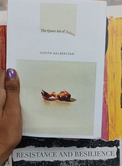 A copy of the book The Queer Art of Failure being held up against a colourful backdrop that has the words 'Resistance and Resilience'. The book is being held up and the image shows the hand of the person holding it. The thumb is painted purple.