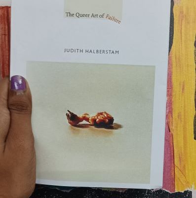 A copy of the book The Queer Art of Failure being held up against a colourful backdrop that has the words 'Resistance and Resilience'. The book is being held up and the image shows the hand of the person holding it. The thumb is painted purple.