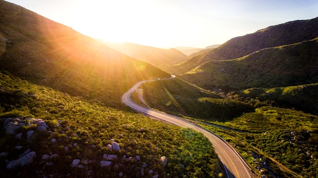 the sun shines behind a row of green hills. a winding road, brown in colour, passes through the hills.