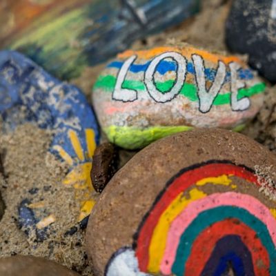 A close-up of vibrantly painted pebbles. One with a rainbow on it is prominently in the foreground. Another with “love” on it is behind it.