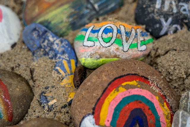 A close-up of vibrantly painted pebbles. One with a rainbow on it is prominently in the foreground. Another with “love” on it is behind it.