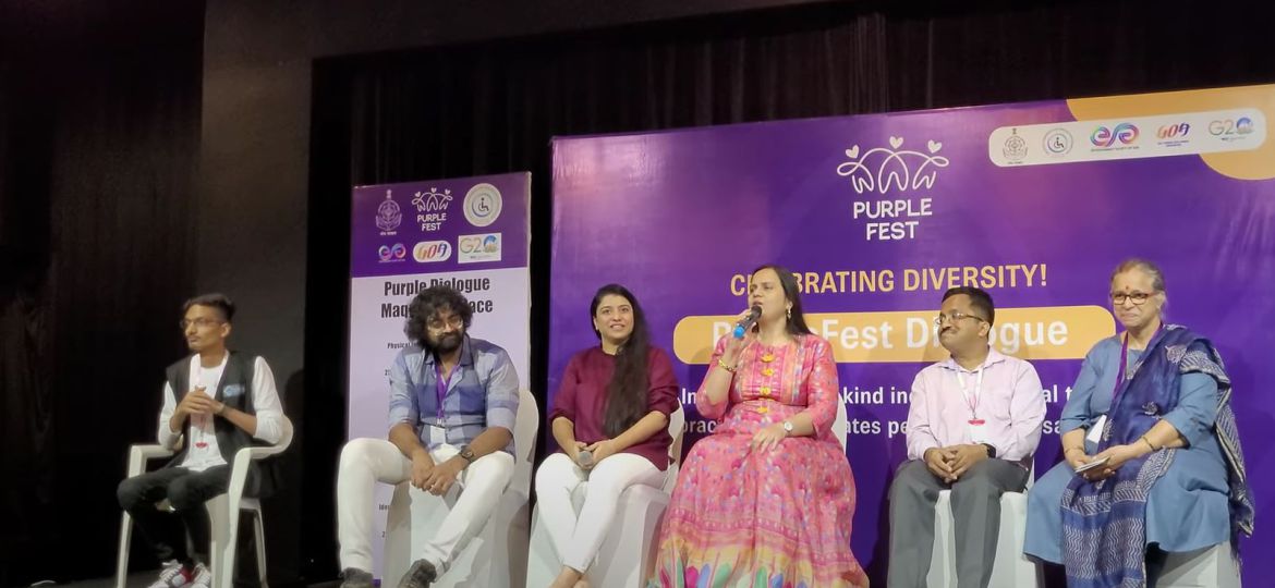 6 people are sitting on a stage. Behind them is a purple poster with the text "Purple Fest" followed by "Celebrating Diversity". The rest of the text is partially hidden. A woman, who is sitting at the centre wearing a pink salwar-kameez, is giving a speech on a mic. The other panellists are looking at her. A man sitting in the far left is translating the speech into sign language.