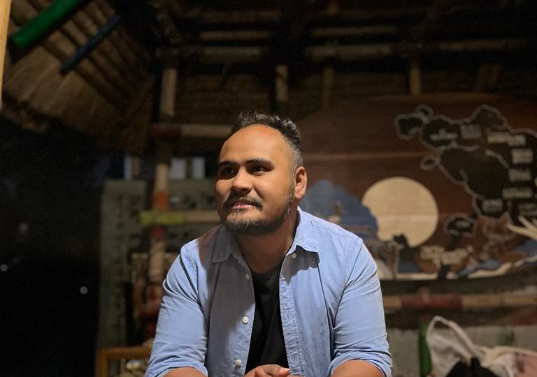 Photograph of Sadam Hanjabam. Sadam has short hair and a moustache. He is a wearing a light blue unbuttoned shirt over a black T-shirt. He is sitting with his hands clasped over a woden table. Behind him is a room with wooden structures and a painting on the wall. A light bulb is illuminating the table.