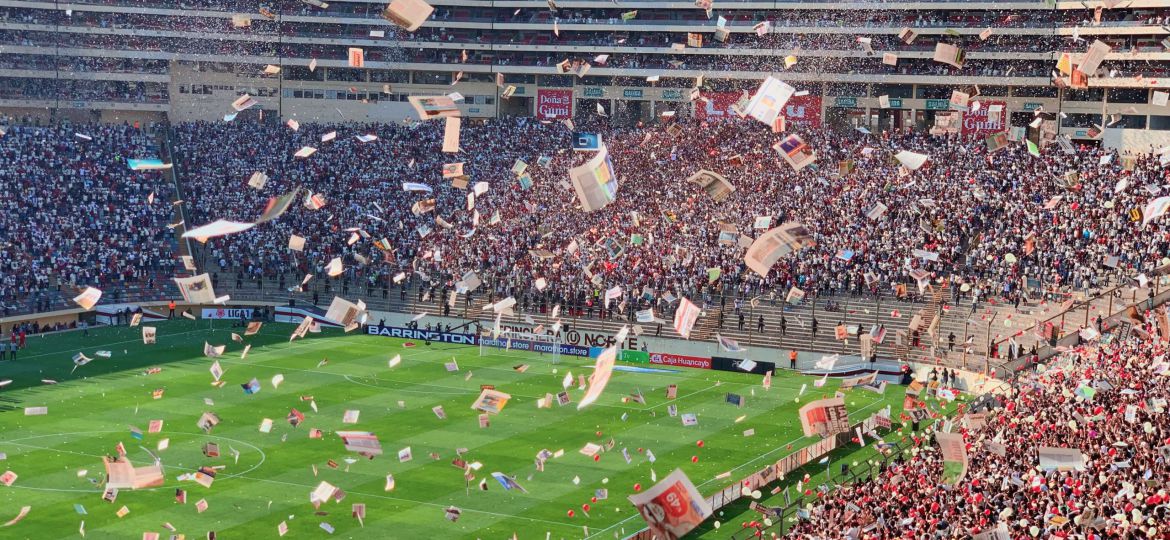 A zoomed-out shot of a football stadium. A lot of pamphlets are flying in the air and one can make out the excitement among the audience.