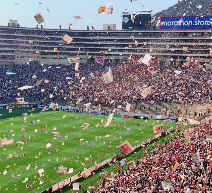 A zoomed-out shot of a football stadium. A lot of pamphlets are flying in the air and one can make out the excitement among the audience.