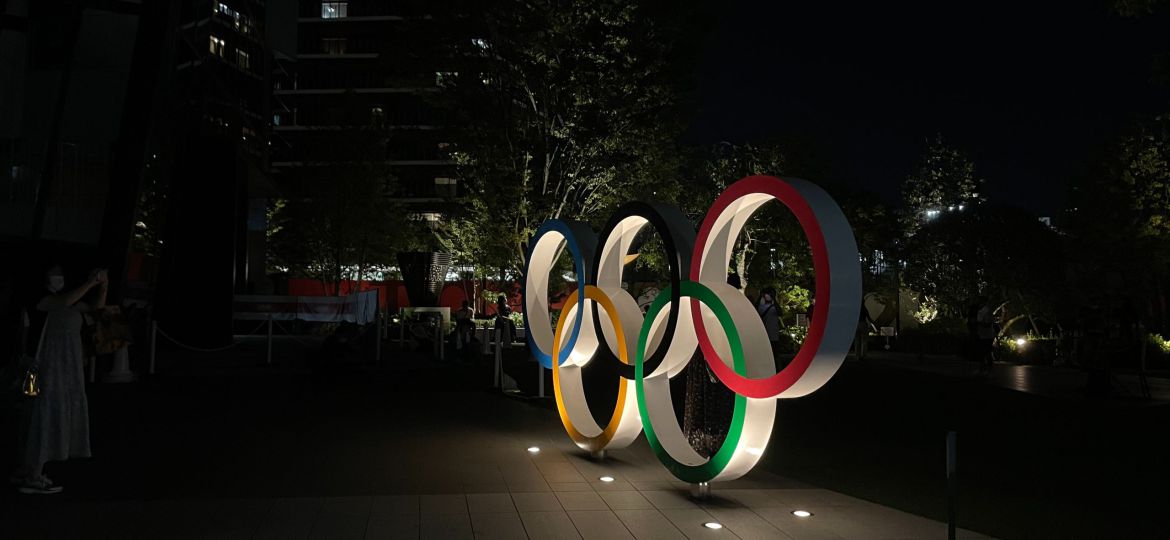 An installation of the Olympic rings in profile towards the right. The time is evening/night and the rings are lit up by lights on the ground.