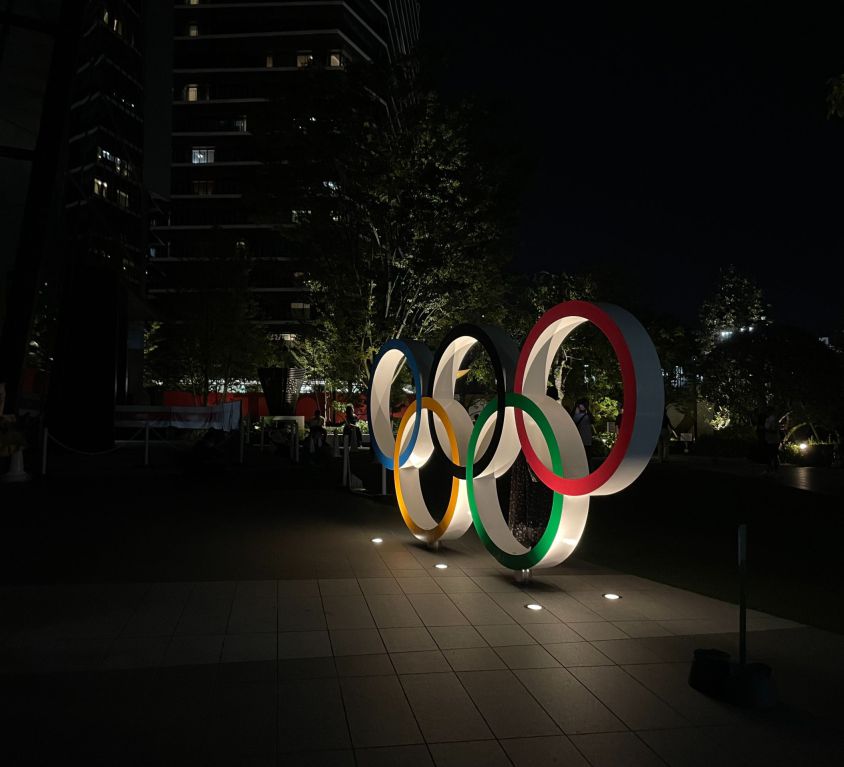 An installation of the Olympic rings in profile towards the right. The time is evening/night and the rings are lit up by lights on the ground.