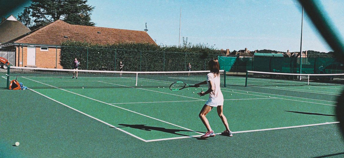 A woman is playing tennis on a court in the distance with her back towards the camera. The shot is captured through the boundary net.