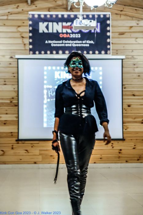 A femme-presenting individual with a short bob and in leather pants, black top, leather waistbelt and leather books walking down a runaway. The individual is wearing a star-shaped mask that covers the top-half of their fact. They are carrying a leather belt in their right hand and is smiling broadly. Behind the individual is a screen and above the screen is a banner that reads "Kink Con Goa 2023: A National Celebration of Kink, Consent and Queerness."