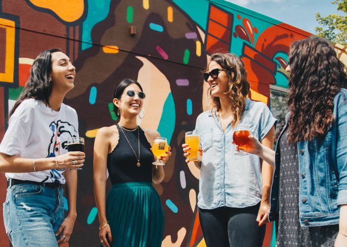 Four femme-presenting individuals standing next to each other against a background of a colourful wall mural in an outdoor setting. Each individual is holding beverages in their hands. They are looking at each other and laughing. 