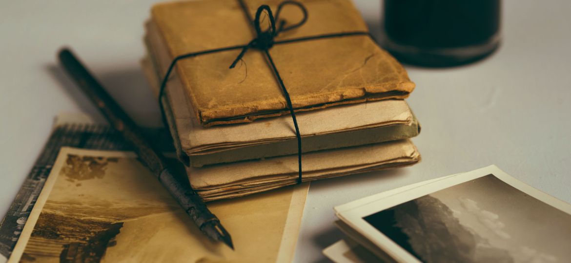 A light-grey surface with faded and yellowed postcards and papers, A small pile of papers and books is tied with string. There is a black fountain pen and an ink bottle is out of focus in one corner.