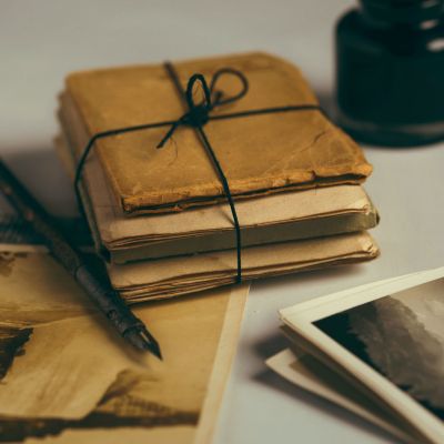 A light-grey surface with faded and yellowed postcards and papers, A small pile of papers and books is tied with string. There is a black fountain pen and an ink bottle is out of focus in one corner.