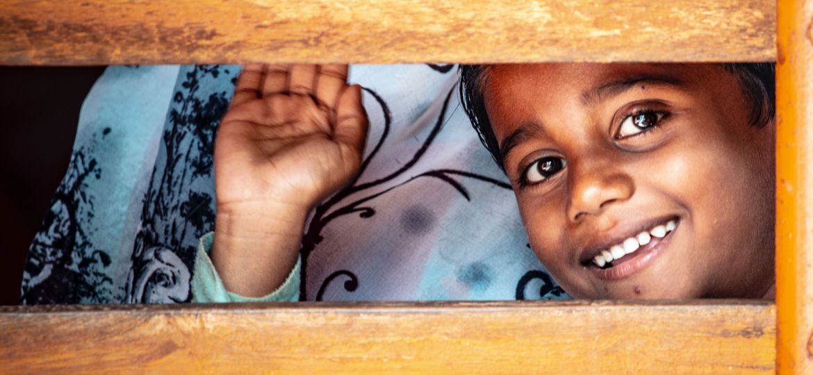 A small boy waves and smilingly peers through a gap between two wooden slats.