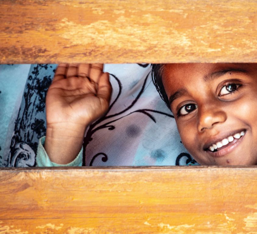 A small boy waves and smilingly peers through a gap between two wooden slats.