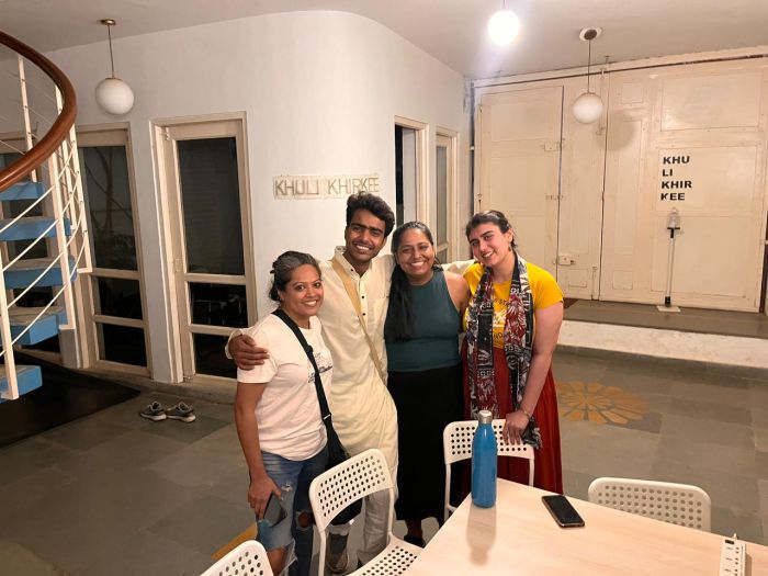 A photograph of four individuals standing behind a white dining set. On the left corner of the image is a staircase bannister. On the wall behind the individuals, there is a banner reading "Khuli Khirkee". The individuals are all smiling and holding one another. 
