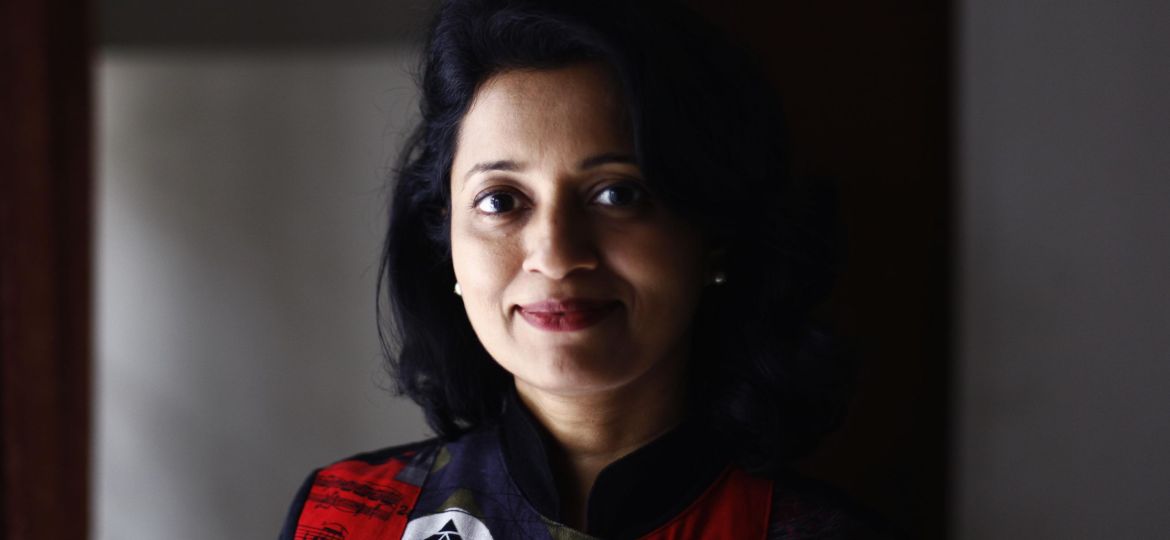 A close-up headshot of Shylashri Shankar. She is against a dark background and half of her face is in shadows. She is smiling and is wearing a red and black checkered shirt with a collar