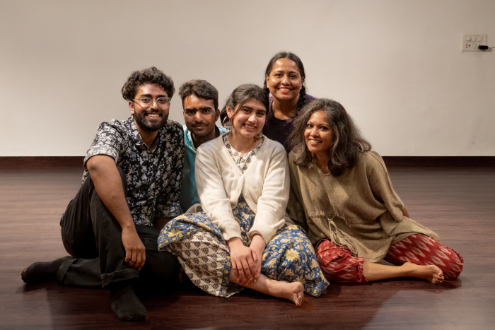 A photograph of the cast and director of the play 'Clay'. There are five individuals sitting on a dark wooden floor, with a beige wall.