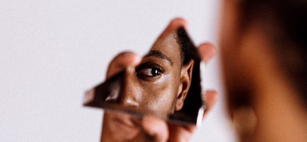 An out-of-focus individual holds a shard of glass in front of them and a portion of their face is visible in it.