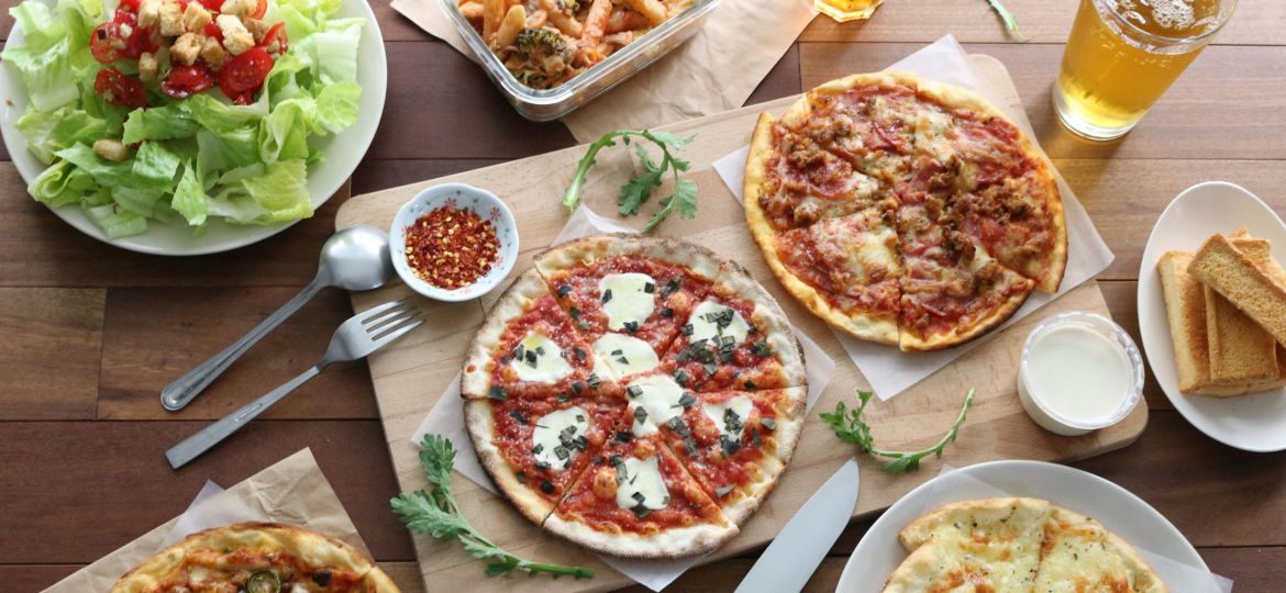 Different kinds of pizzas, a caesar salad, pasta, bread, dips, and drinks kept on a wooden surface.