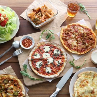 Different kinds of pizzas, a caesar salad, pasta, bread, dips, and drinks kept on a wooden surface.