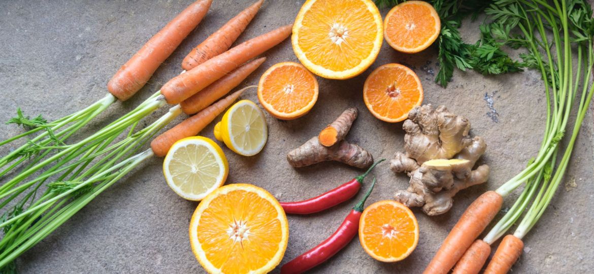 Sliced oranges and lemons along with whole carrots, ginger, turmeric, and red chillies on a rough grey surface.