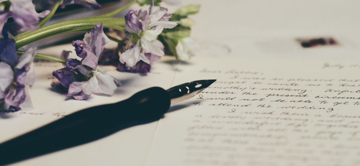 A close-up photograph of a white blank paper upon which letters are scribbled. Upon the paper is a black fountain pen and some violet flowers.