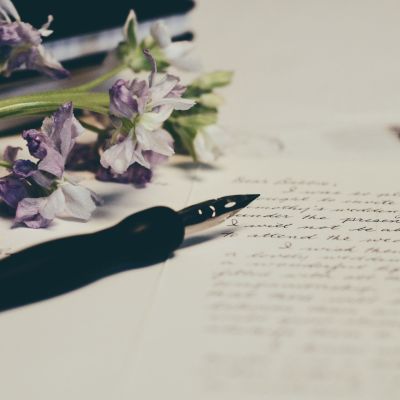 A close-up photograph of a white blank paper upon which letters are scribbled. Upon the paper is a black fountain pen and some violet flowers.