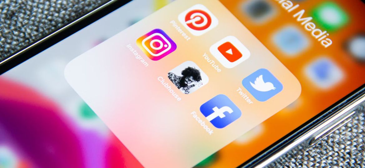 Close-up of a phone screen with a menu folder titled "Social Media" open. Icons of Pinterest, YouTube, Twitter, Instagram, Clubhouse, and Facebook are visible.