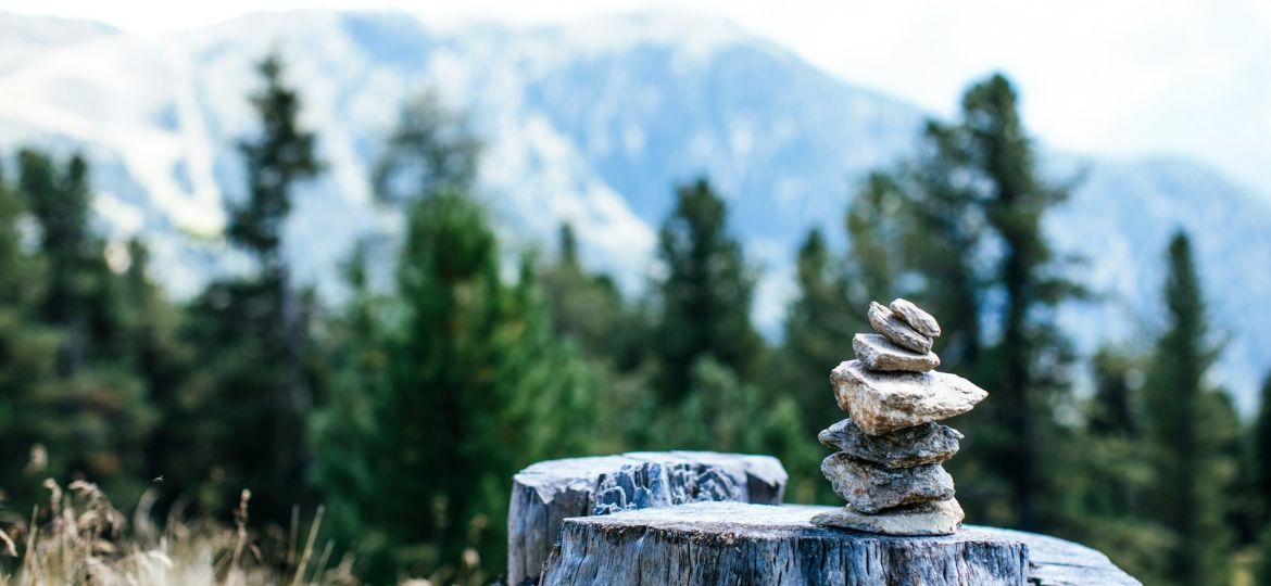 a pile of little stones are stacked vertically on a rock. in the background are green trees and snowy mountains