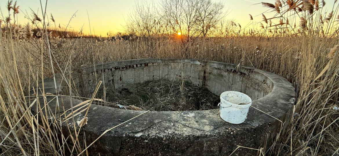 A small white plastic bucket sitting on top of a dried up stone well in a field with the sun setting on the horizon. The landscape is very dry.