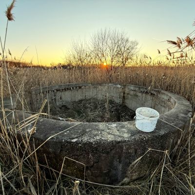A small white plastic bucket sitting on top of a dried up stone well in a field with the sun setting on the horizon. The landscape is very dry.