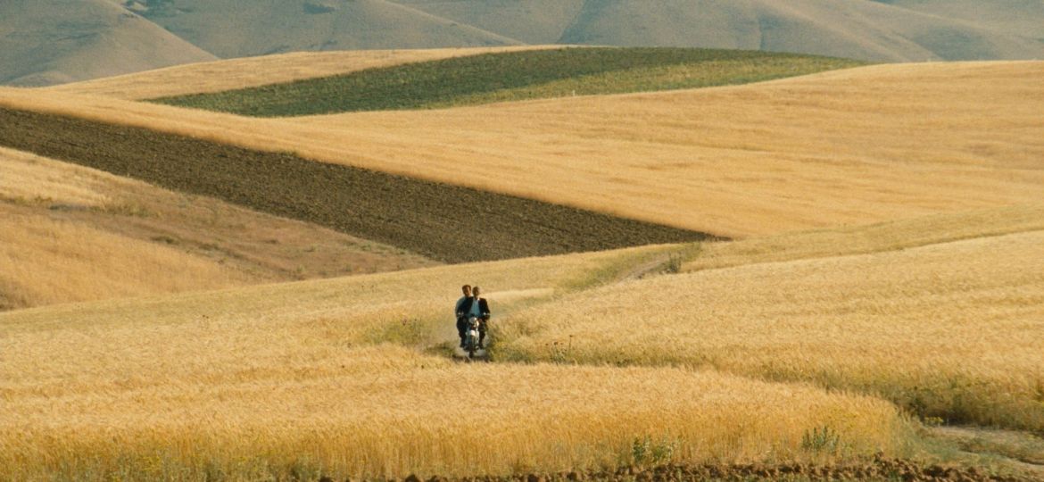 A still from the film The wind will carry us. The image is of a yellow field landscape with hills at the background and a motor bike in the centre