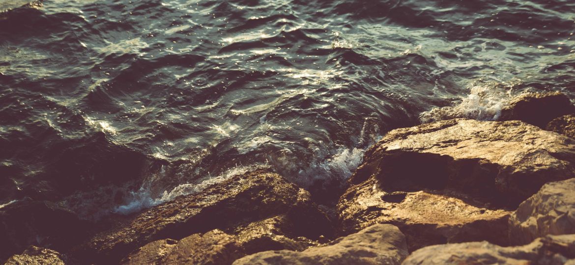 Mild waves crash on rocks, where the surfaces of both the water and rocks reflect sunlight