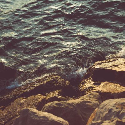 Mild waves crash on rocks, where the surfaces of both the water and rocks reflect sunlight