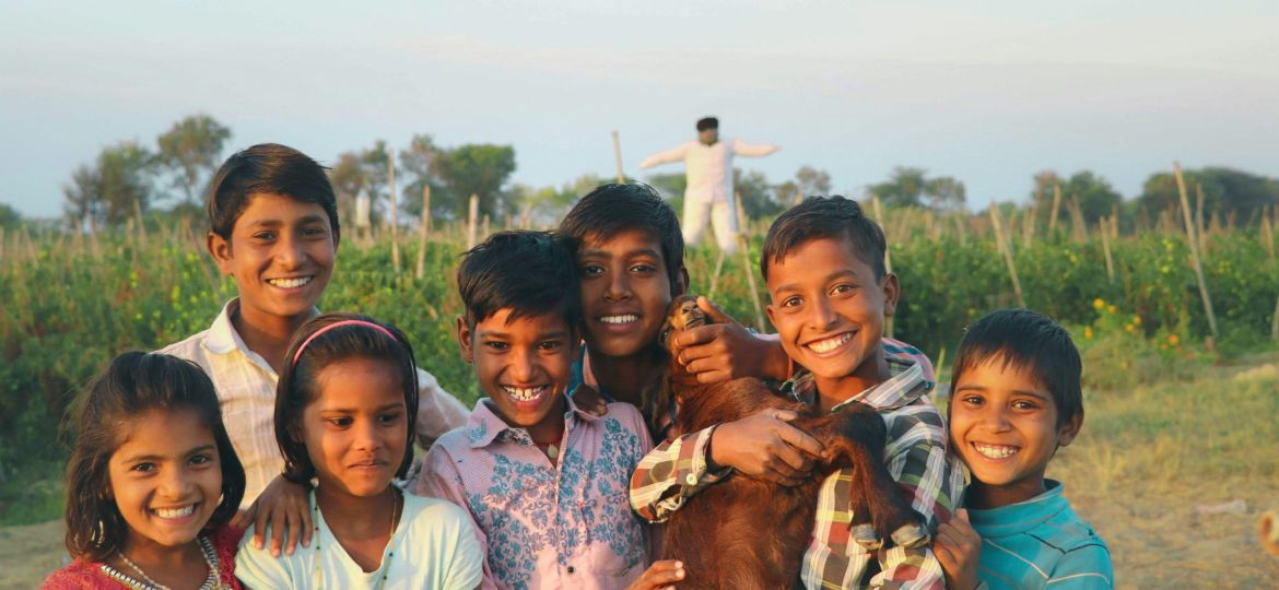 A group of children posing for the camera with smiling faces with the backdrop of a field with a scarecrow. One of the boys is holding a goat