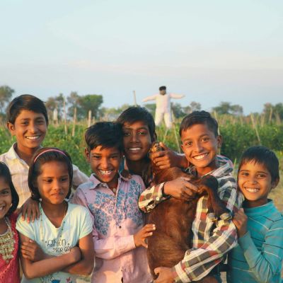 A group of children posing for the camera with smiling faces with the backdrop of a field with a scarecrow. One of the boys is holding a goat