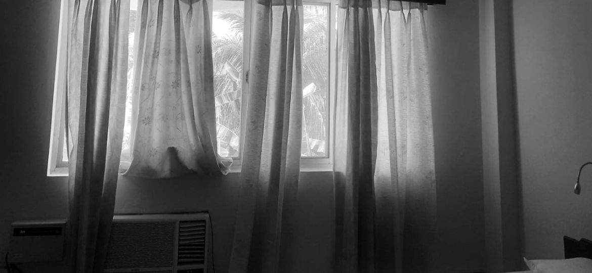 A black & white photo of a neatly made bed along with sheer curtains drawn over the window in the room depicts a quiet personal space