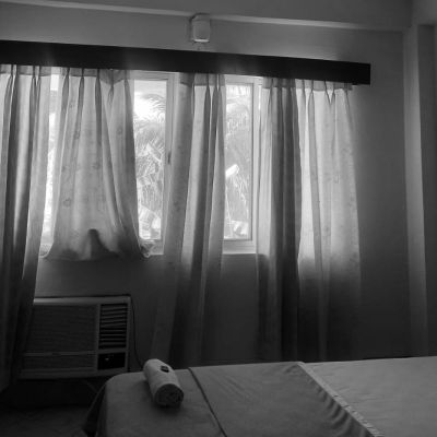 A black & white photo of a neatly made bed along with sheer curtains drawn over the window in the room depicts a quiet personal space