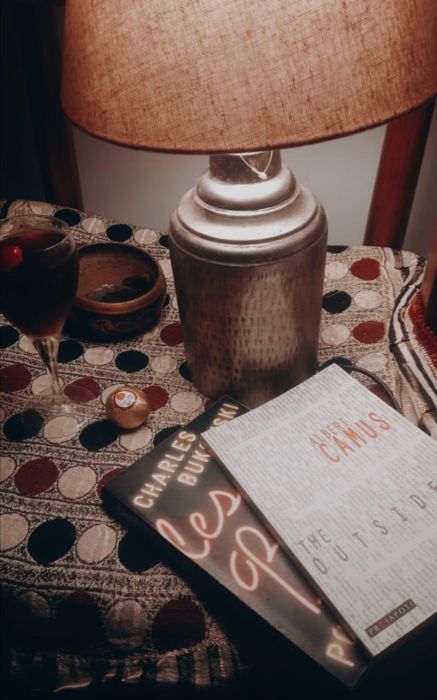 a table top with a glass of wine, a table lamp and two books: Alber Camus The Outsider and Charles Bukowski's Post Office